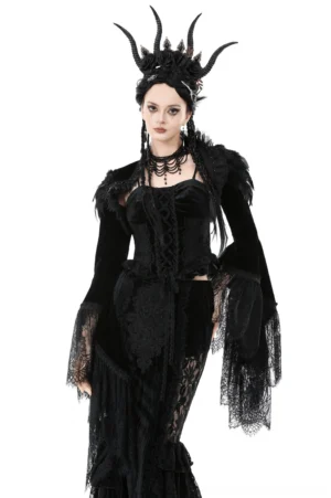 Front three-quarter view of black velvet gothic shrug with feathered shoulders, lace bell sleeves, and ruffled details worn by model with horned feather headdress on white studio background