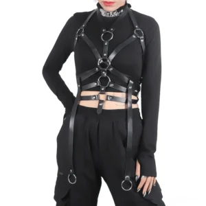 Black synthetic leather bondage chest harness with metal rings and adjustable straps worn over fitted long-sleeve crop top on model with tattooed arms