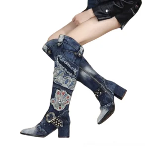 Dark blue denim gothic knee-high boots worn on female model, side view displaying studded rivets, embroidered patches, and block heel silhouette