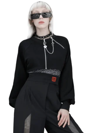 Black chain-adorned crop jacket with dual front zippers and heart charm detail, displayed on platinum blonde model in studio setting