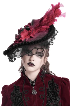 Female model wearing burgundy and black gothic fascinator hat with lace veil and feather accents, front portrait view showcasing dramatic makeup and dark aesthetic
