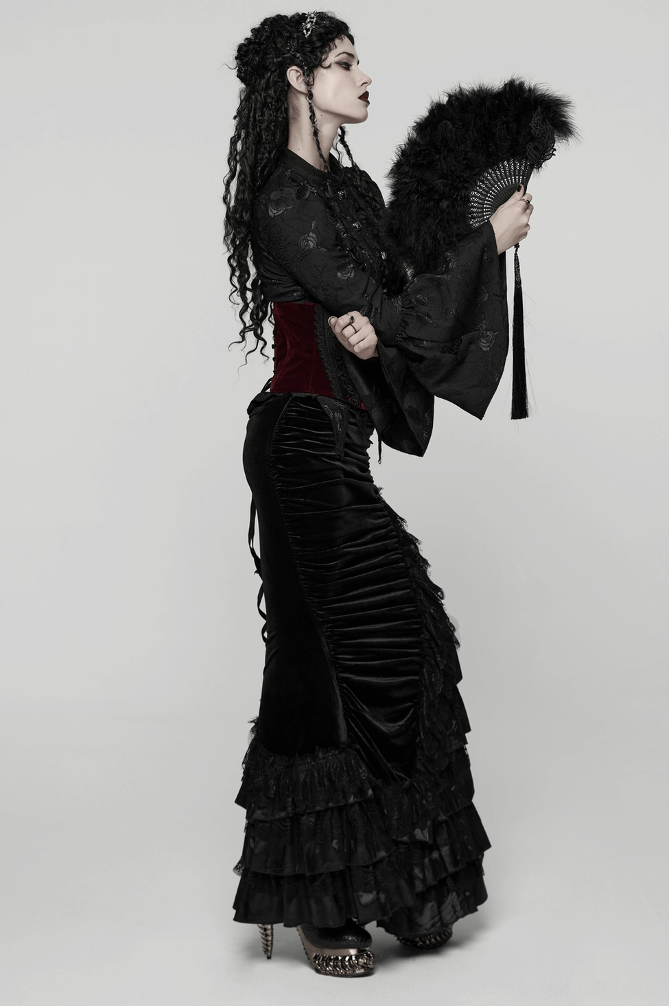 Right profile of model with curled dark hair holding gothic black feather fan, wearing velvet ruffled skirt
