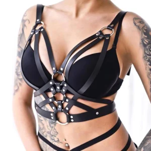 Black PU leather gothic harness bra top with crisscross straps and metal O-ring accents, front close-up view on tattooed model showing adjustable lace-up design