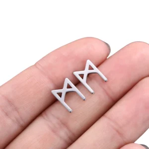 Silver gothic M-shaped rune stud earring held in fingertips, close-up overhead view showcasing hollow stainless steel design with polished finish