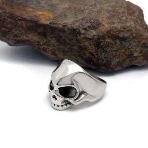 Alternative fashion skull ring left side profile view displayed against rough black rock, showing smooth polished silver stainless steel construction and simplified skull silhouette design