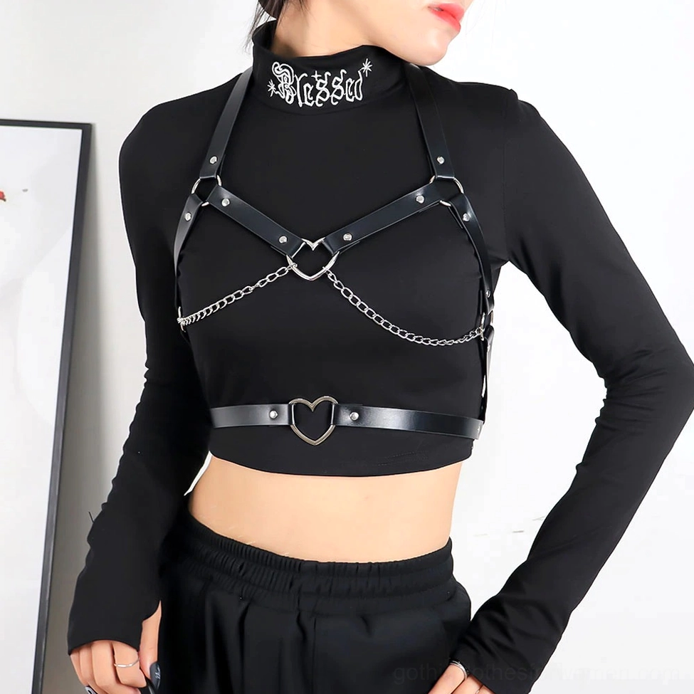 Female model wearing black leather chest harness over crop top with silver chain and heart hardware, indoor setting