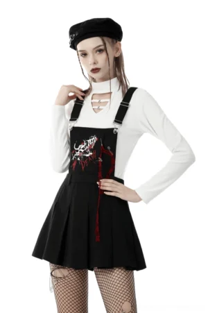 Black skeleton pleated pinafore dress with adjustable straps, front three-quarter view showing embroidered skull design with red tassel trim