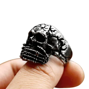 Vintage stainless steel skull biker ring worn on male thumb, front view showcasing skeletal hand motif and weathered antique-finish texture