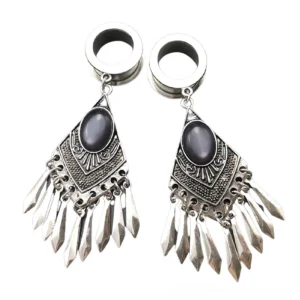 Tribal gothic tunnel earrings pair - silver surgical steel with dark oval gemstones and carved tribal patterns with spike tassel pendants, flat lay overhead view